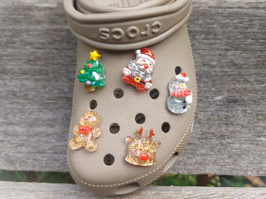 A glittery Christmas-themed set of five shoe charms featuring a gingerbread man, reindeer, snowman, Santa, and a Christmas tree. Each charm has a sparkling glitter finish, adding a festive and playful holiday touch to your shoes.
