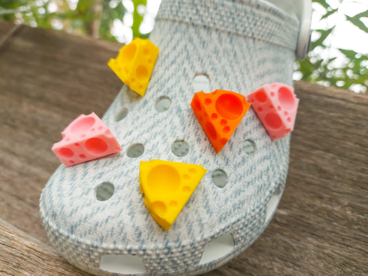 Close-up of a pair of light blue shoes decorated with colorful 3D-printed cheese wedge charms in shades of yellow, orange, and pink. The cheese charms feature realistic holes and a playful, quirky design, adding a fun pop of color to the footwear.