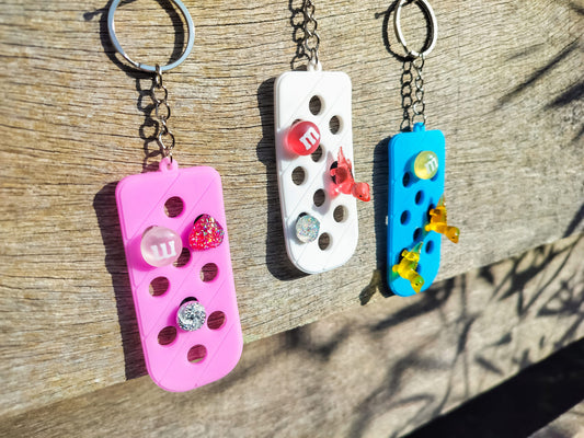 Three PVC keychains in pink, white, and blue with holes designed to hold shoe charms. Each keychain displays a unique set of charms, including candy-themed and colorful glitter charms, showcasing a playful and customizable accessory for charm lovers.