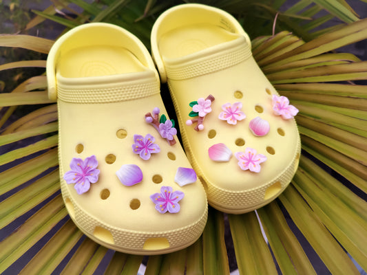 A pair of pale yellow Croc-style clogs decorated with pink and purple flower charms, placed on green palm leaves.
