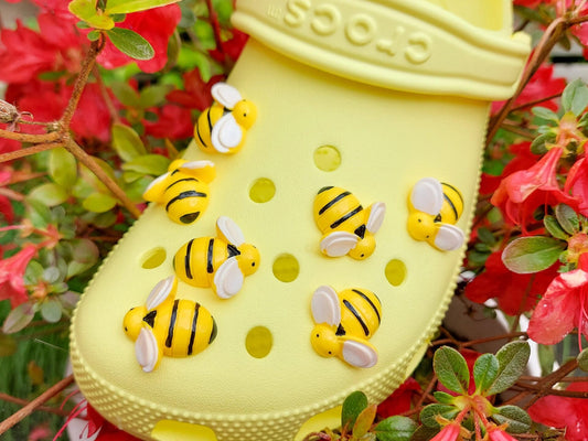 A yellow Croc-style clog decorated with multiple yellow and black bumblebee charms, set against a background of red flowers and green foliage.