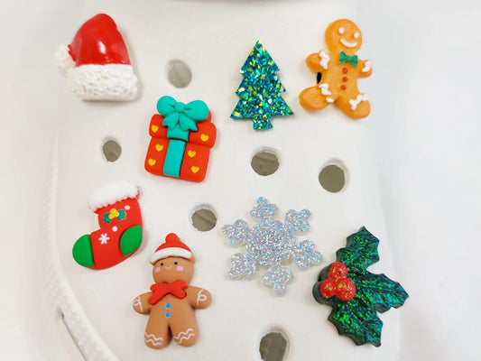 A set of eight Christmas-themed clog charms displayed on a white clog shoe. The charms include a red Santa hat, a green glitter tree, a gingerbread man, a red gift box with a teal bow, a red and green stocking, a silver glitter snowflake, a green holly leaf with red berries, and a brown gingerbread man.