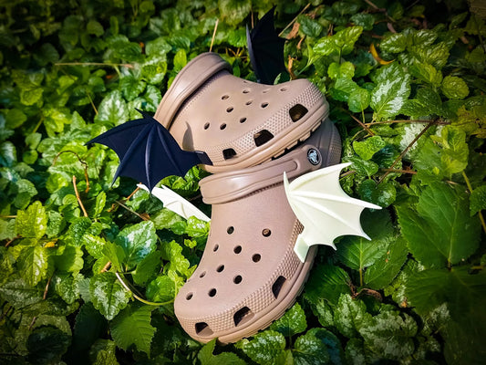 A pair of tan Croc-style clogs placed on dense green foliage, with small black and white bat wings attached near the shoes.