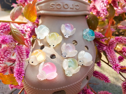 Close-up of glow-in-the-dark firefly charms on beige shoes, surrounded by vibrant pink flowers. The charming fireflies light up in different colors, creating a magical and whimsical look, perfect for adding a soft, glowing touch to your footwear.