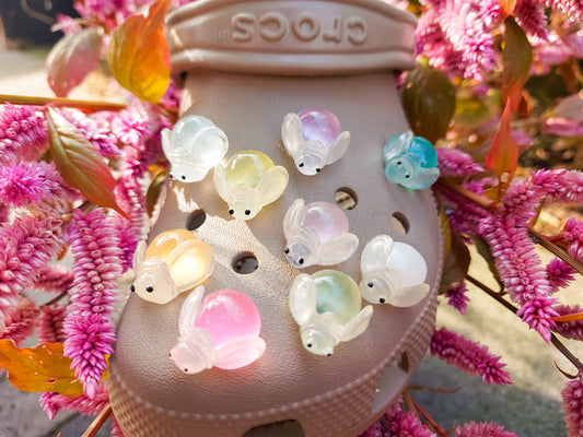 Close-up of glow-in-the-dark firefly charms on beige shoes, surrounded by vibrant pink flowers. The charming fireflies light up in different colors, creating a magical and whimsical look, perfect for adding a soft, glowing touch to your footwear.