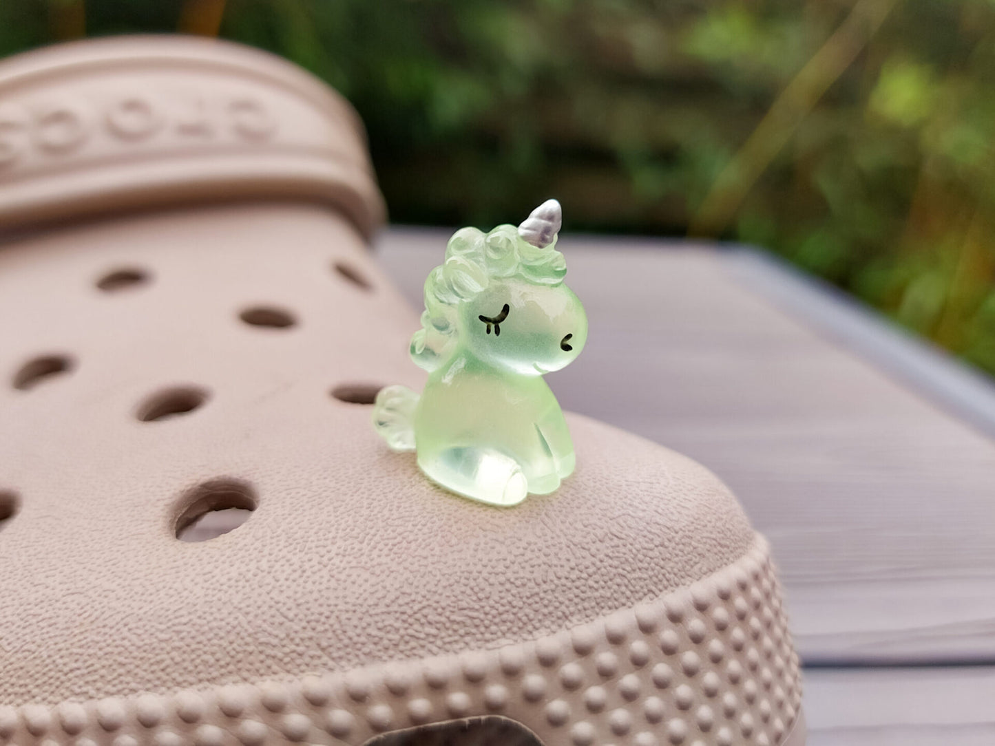 Beige shoes decorated with glow-in-the-dark unicorn charms in various pastel colors, including purple, green, pink, orange, yellow, and blue, placed on grass. The charms add a whimsical and magical touch to the shoes.