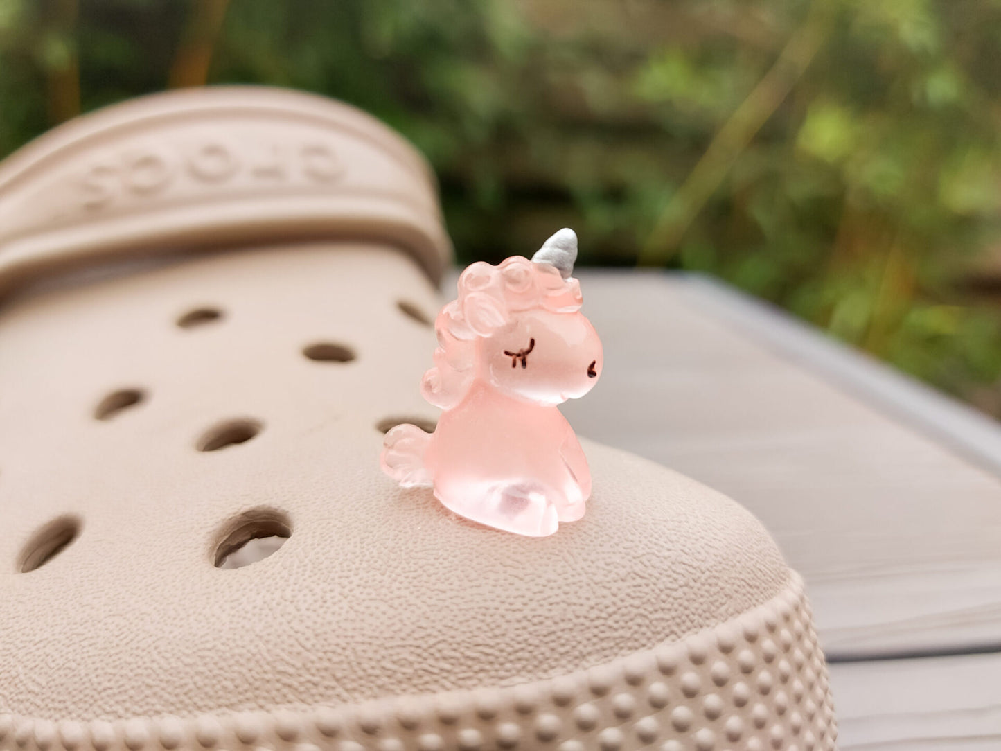 Beige shoes decorated with glow-in-the-dark unicorn charms in various pastel colors, including purple, green, pink, orange, yellow, and blue, placed on grass. The charms add a whimsical and magical touch to the shoes.