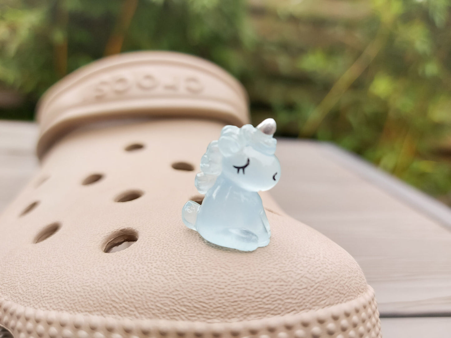 Beige shoes decorated with glow-in-the-dark unicorn charms in various pastel colors, including purple, green, pink, orange, yellow, and blue, placed on grass. The charms add a whimsical and magical touch to the shoes.