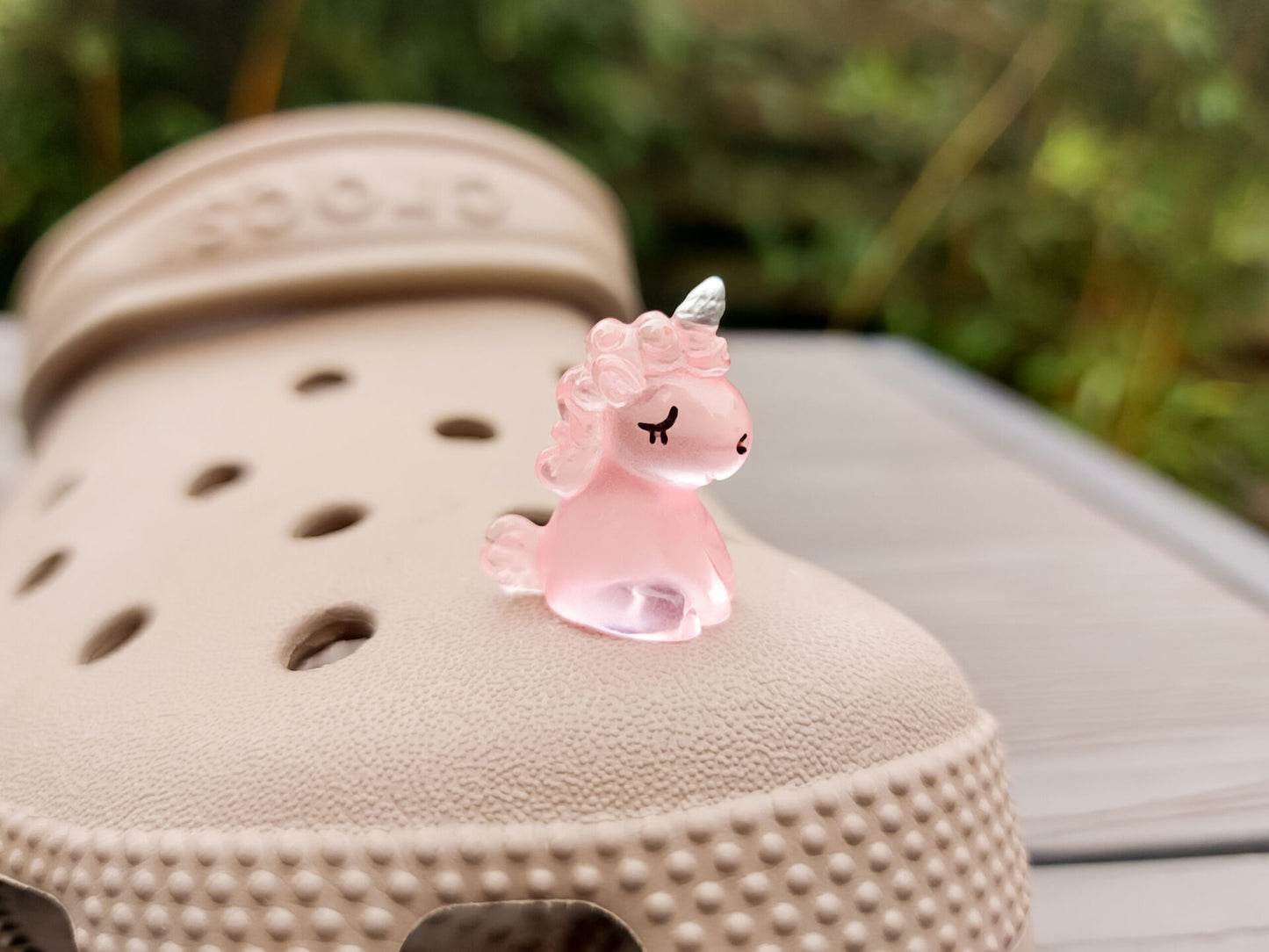 Beige shoes decorated with glow-in-the-dark unicorn charms in various pastel colors, including purple, green, pink, orange, yellow, and blue, placed on grass. The charms add a whimsical and magical touch to the shoes.