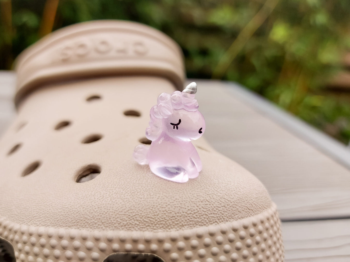 Beige shoes decorated with glow-in-the-dark unicorn charms in various pastel colors, including purple, green, pink, orange, yellow, and blue, placed on grass. The charms add a whimsical and magical touch to the shoes.