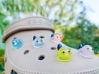 Taupe-colored shoe shoe adorned with glow-in-the-dark bird charms. Each charm features a small, round bird with a beak and black wings, glowing softly in various colors. The shoe is positioned outdoors against a backdrop of green foliage.
