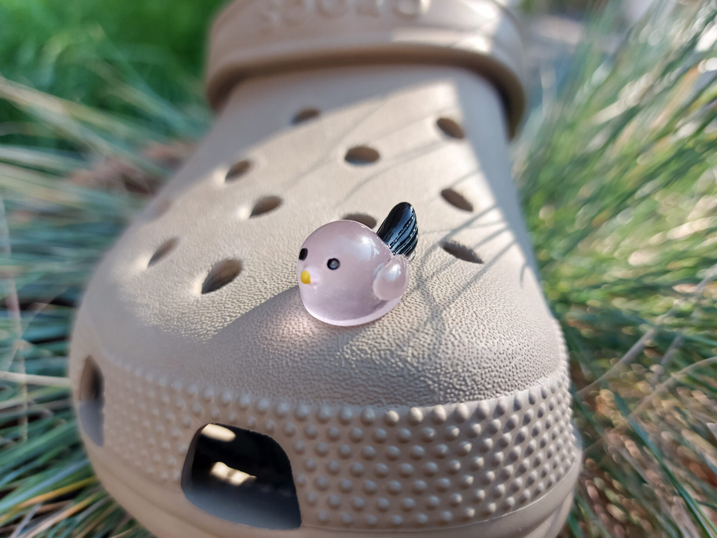 Taupe-colored shoe shoe adorned with glow-in-the-dark bird charms. Each charm features a small, round bird with a beak and black wings, glowing softly in various colors. The shoe is positioned outdoors against a backdrop of green foliage.