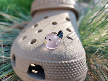 Taupe-colored shoe shoe adorned with glow-in-the-dark bird charms. Each charm features a small, round bird with a beak and black wings, glowing softly in various colors. The shoe is positioned outdoors against a backdrop of green foliage.
