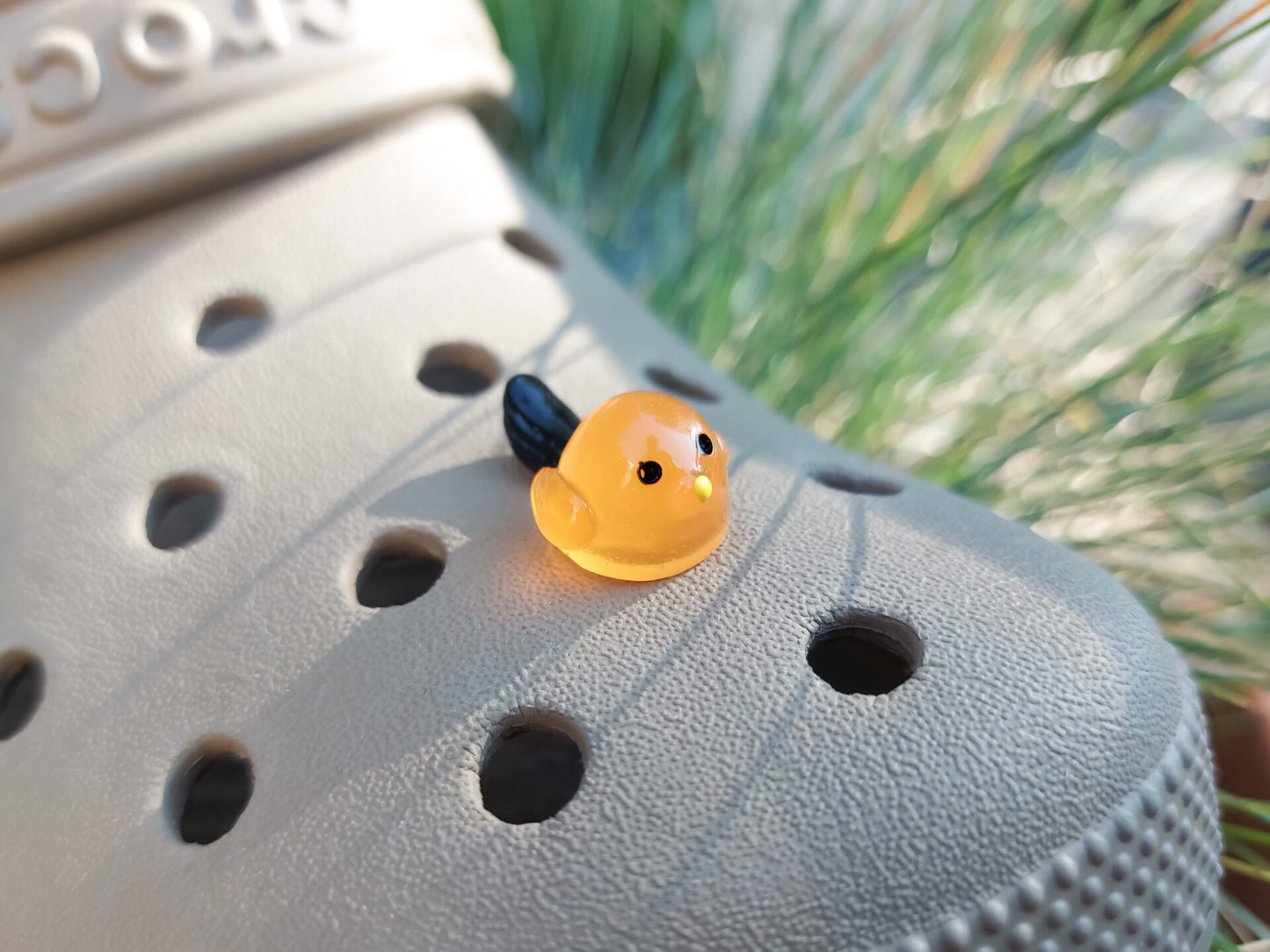 Taupe-colored shoe shoe adorned with glow-in-the-dark bird charms. Each charm features a small, round bird with a beak and black wings, glowing softly in various colors. The shoe is positioned outdoors against a backdrop of green foliage.