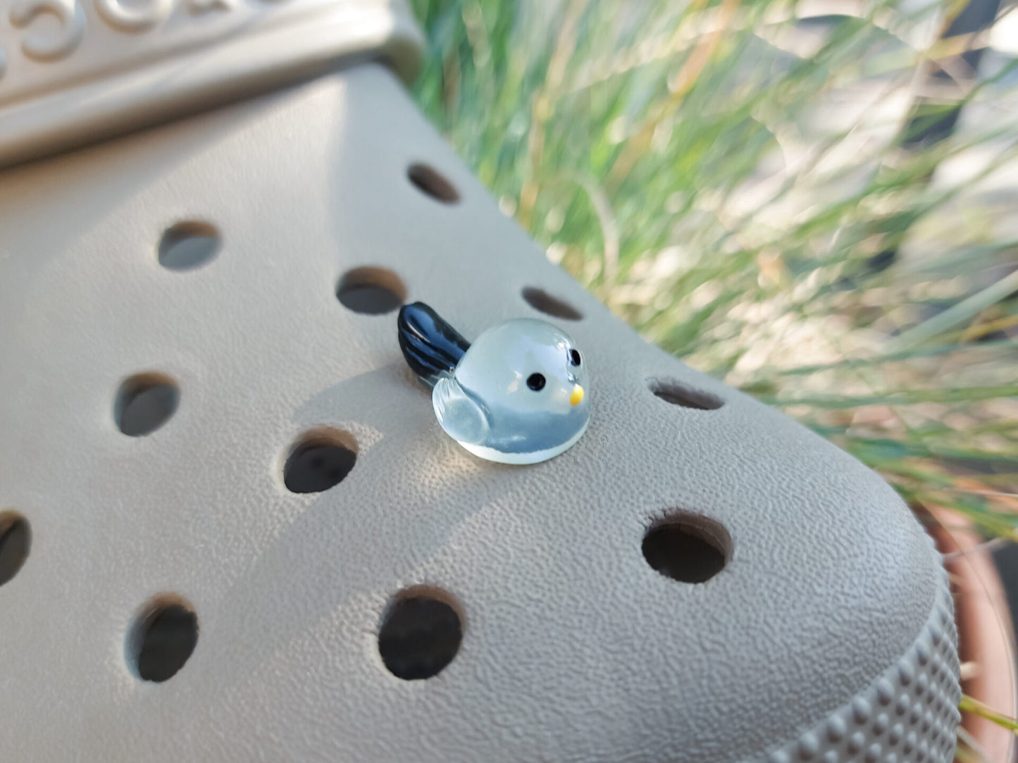 Taupe-colored shoe shoe adorned with glow-in-the-dark bird charms. Each charm features a small, round bird with a beak and black wings, glowing softly in various colors. The shoe is positioned outdoors against a backdrop of green foliage.
