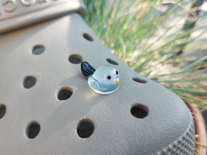 Taupe-colored shoe shoe adorned with glow-in-the-dark bird charms. Each charm features a small, round bird with a beak and black wings, glowing softly in various colors. The shoe is positioned outdoors against a backdrop of green foliage.