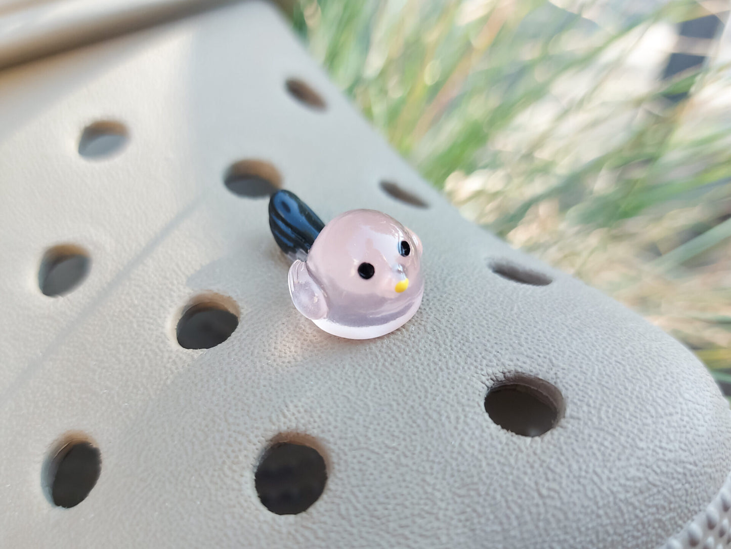 Taupe-colored shoe shoe adorned with glow-in-the-dark bird charms. Each charm features a small, round bird with a beak and black wings, glowing softly in various colors. The shoe is positioned outdoors against a backdrop of green foliage.