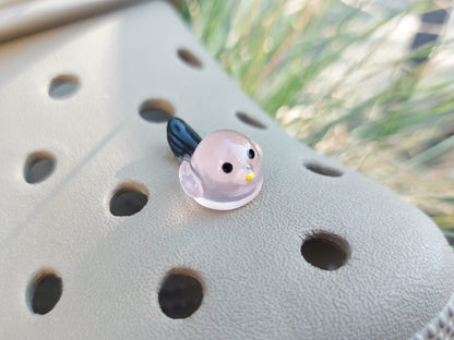 Taupe-colored shoe shoe adorned with glow-in-the-dark bird charms. Each charm features a small, round bird with a beak and black wings, glowing softly in various colors. The shoe is positioned outdoors against a backdrop of green foliage.