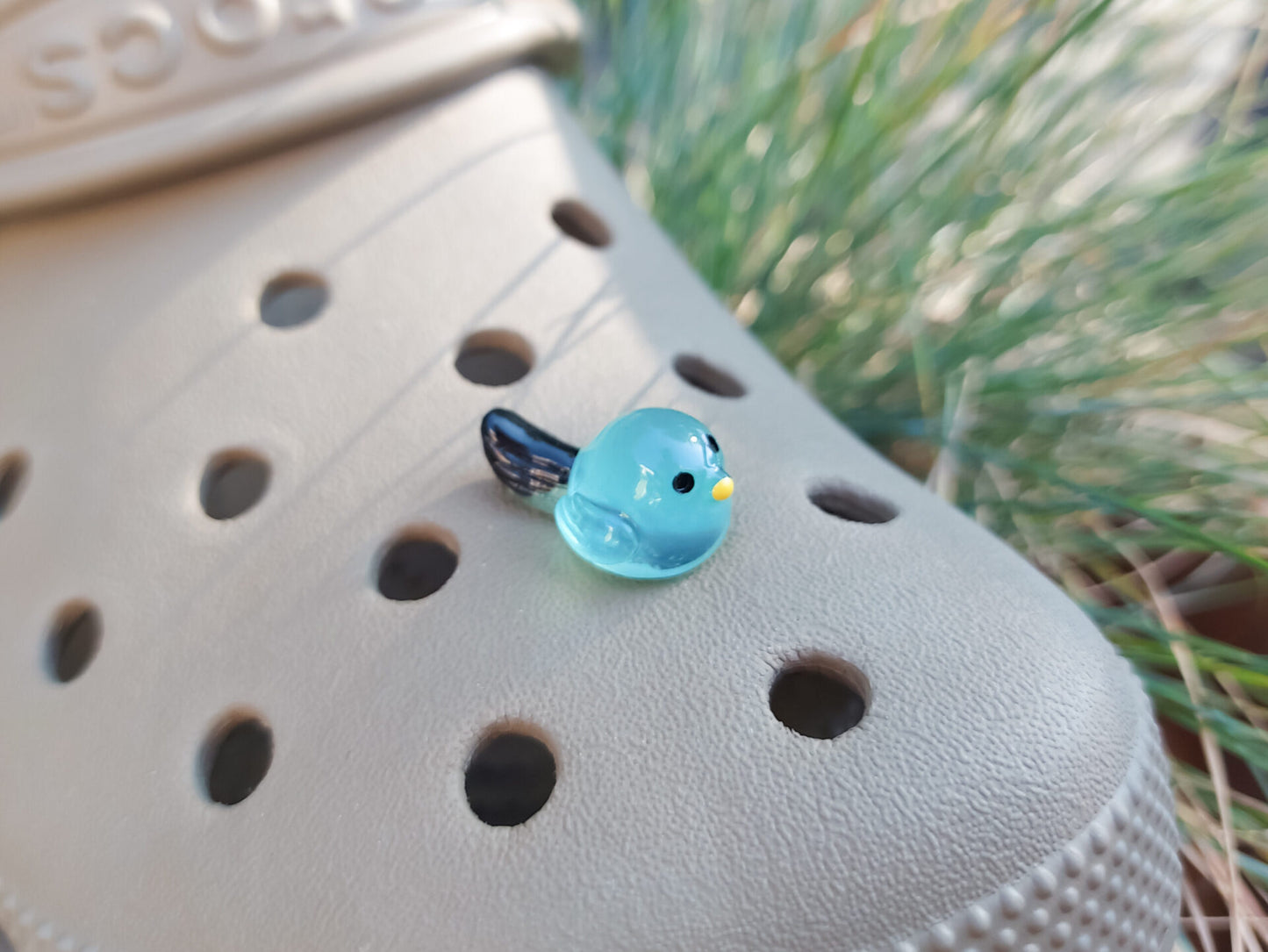 Taupe-colored shoe shoe adorned with glow-in-the-dark bird charms. Each charm features a small, round bird with a beak and black wings, glowing softly in various colors. The shoe is positioned outdoors against a backdrop of green foliage.