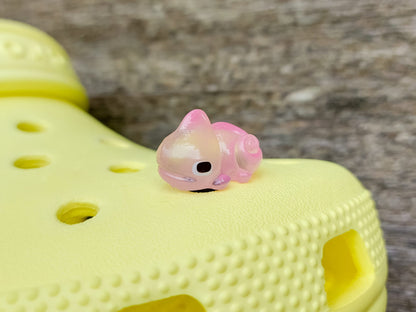 A close-up of a yellow shoe shoe with vibrant glow-in-the-dark chameleon charms in different colors (green, orange, pink, blue, and yellow) displayed outdoors on green grass.