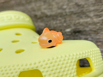 A close-up of a yellow shoe shoe with vibrant glow-in-the-dark chameleon charms in different colors (green, orange, pink, blue, and yellow) displayed outdoors on green grass.