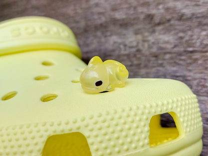 A close-up of a yellow shoe shoe with vibrant glow-in-the-dark chameleon charms in different colors (green, orange, pink, blue, and yellow) displayed outdoors on green grass.