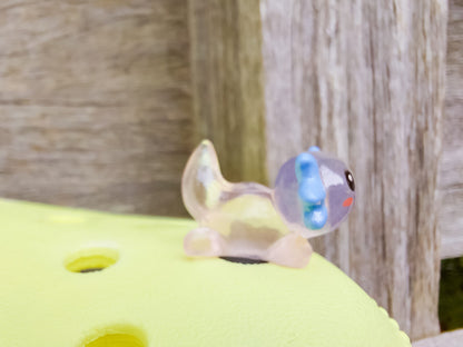 A yellow shoe shoe featuring colorful glow-in-the-dark axolotl charms in shades of green, pink, blue, orange, and silver, displayed outdoors on a wooden surface. The charms, with smiling faces and frilly gills, brighten the shoe.