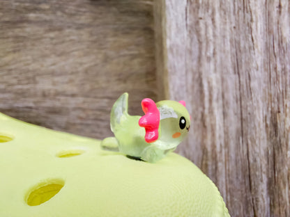 A yellow shoe shoe featuring colorful glow-in-the-dark axolotl charms in shades of green, pink, blue, orange, and silver, displayed outdoors on a wooden surface. The charms, with smiling faces and frilly gills, brighten the shoe.