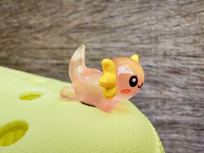 A yellow shoe shoe featuring colorful glow-in-the-dark axolotl charms in shades of green, pink, blue, orange, and silver, displayed outdoors on a wooden surface. The charms, with smiling faces and frilly gills, brighten the shoe.