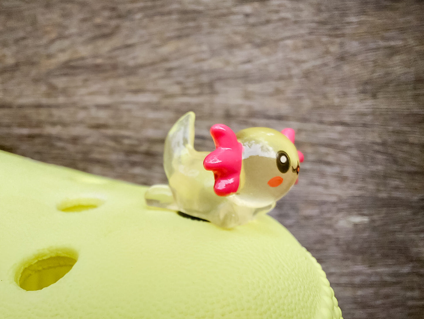 A yellow shoe shoe featuring colorful glow-in-the-dark axolotl charms in shades of green, pink, blue, orange, and silver, displayed outdoors on a wooden surface. The charms, with smiling faces and frilly gills, brighten the shoe.