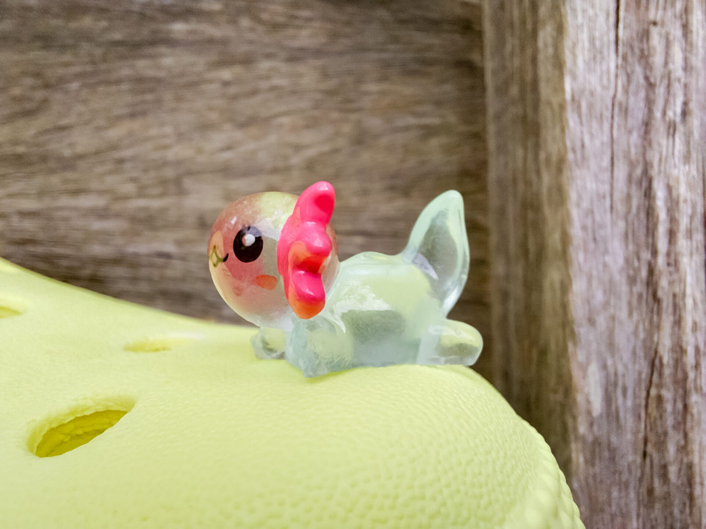 A yellow shoe shoe featuring colorful glow-in-the-dark axolotl charms in shades of green, pink, blue, orange, and silver, displayed outdoors on a wooden surface. The charms, with smiling faces and frilly gills, brighten the shoe.