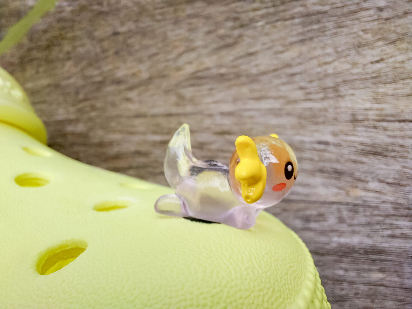 A yellow shoe shoe featuring colorful glow-in-the-dark axolotl charms in shades of green, pink, blue, orange, and silver, displayed outdoors on a wooden surface. The charms, with smiling faces and frilly gills, brighten the shoe.