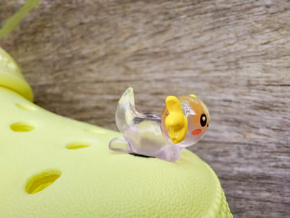 A yellow shoe shoe featuring colorful glow-in-the-dark axolotl charms in shades of green, pink, blue, orange, and silver, displayed outdoors on a wooden surface. The charms, with smiling faces and frilly gills, brighten the shoe.