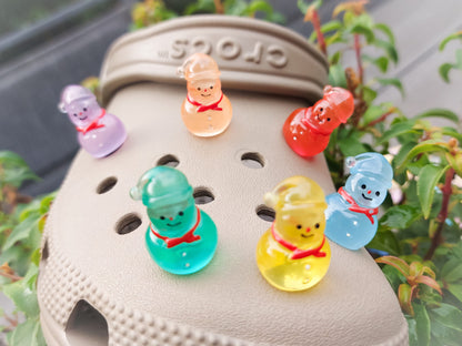 Glow-in-the-dark snowmen charms adorning a beige shoes shoe. The snowmen are in vibrant colors like green, yellow, red, purple, orange, and blue. Each snowman has a scarf, and a small hat, radiating a festive vibe perfect for the holiday season.