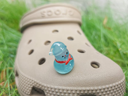 Glow-in-the-dark snowmen charms adorning a beige shoes shoe. The snowmen are in vibrant colors like green, yellow, red, purple, orange, and blue. Each snowman has a scarf, and a small hat, radiating a festive vibe perfect for the holiday season.