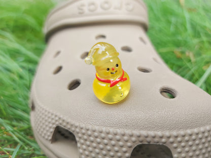 Glow-in-the-dark snowmen charms adorning a beige shoes shoe. The snowmen are in vibrant colors like green, yellow, red, purple, orange, and blue. Each snowman has a scarf, and a small hat, radiating a festive vibe perfect for the holiday season.