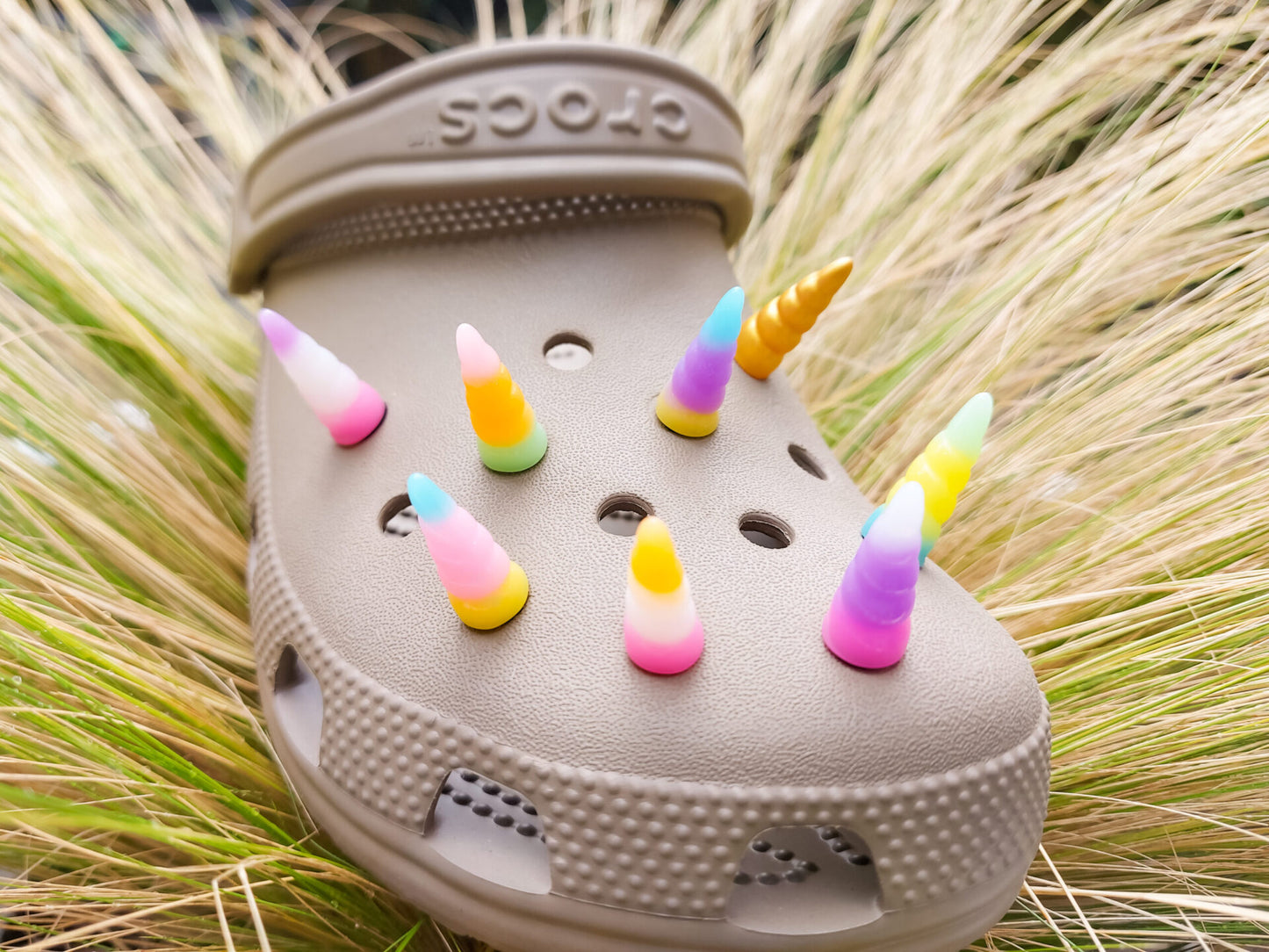 A tan shoes shoe decorated with several colorful unicorn horn charms, each one blending from one pastel shade to another in shades of pink, yellow, blue, and purple. The shoe is displayed on a backdrop of light green grasses.