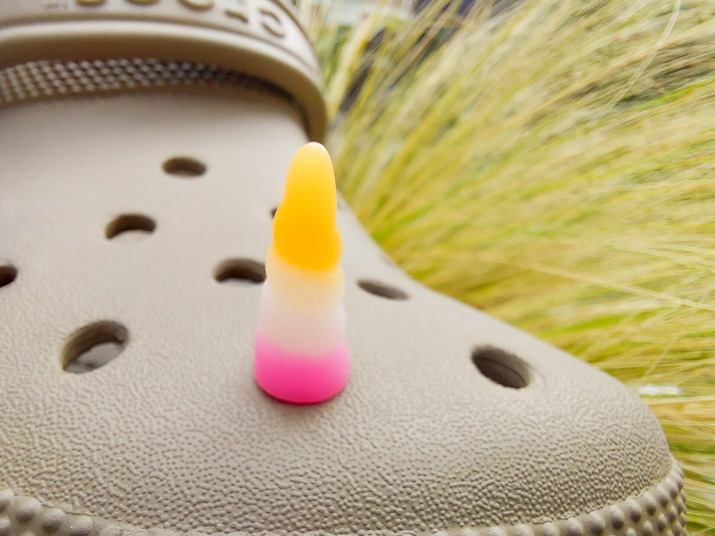 A tan shoes shoe decorated with several colorful unicorn horn charms, each one blending from one pastel shade to another in shades of pink, yellow, blue, and purple. The shoe is displayed on a backdrop of light green grasses.