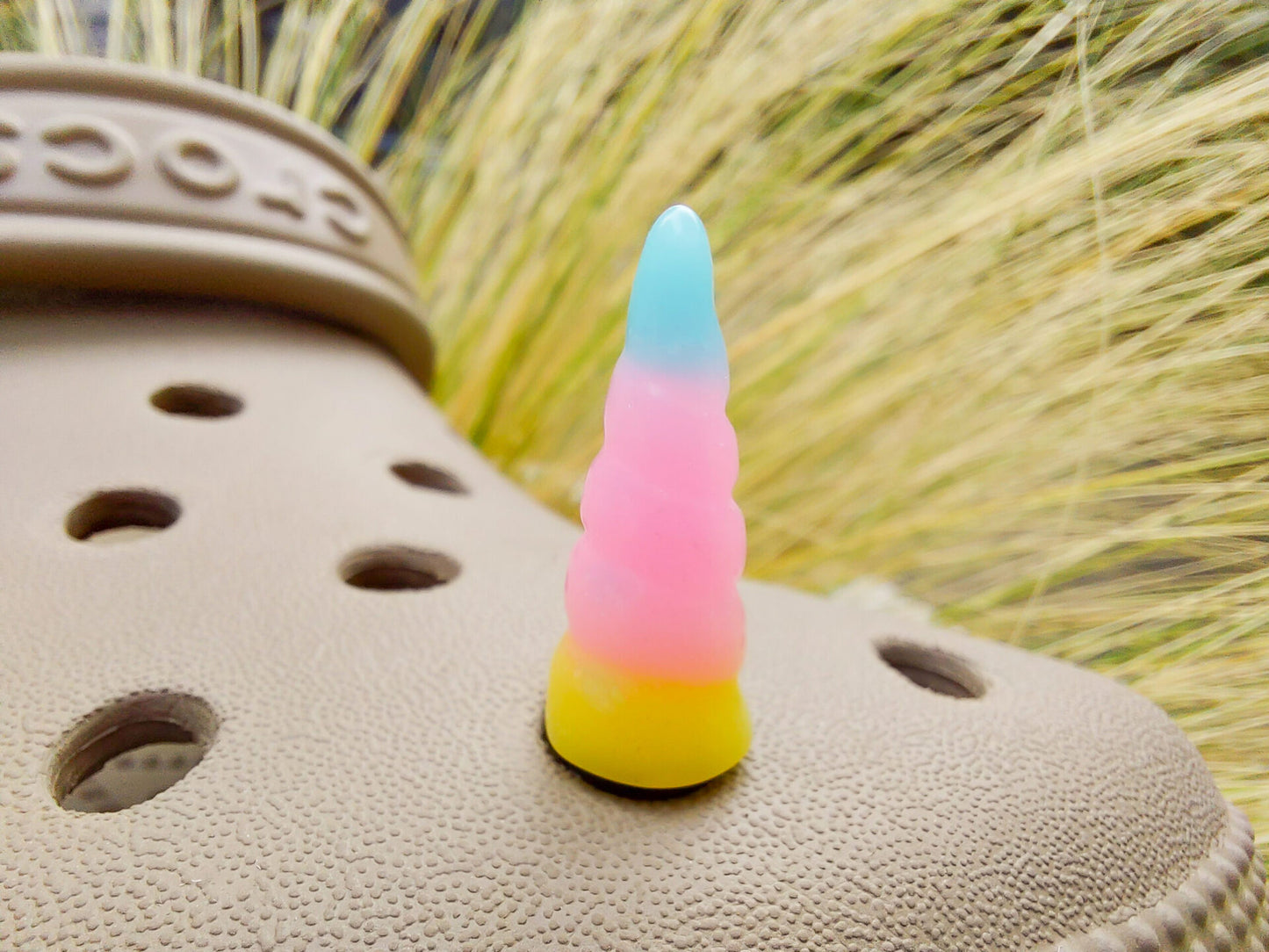 A tan shoes shoe decorated with several colorful unicorn horn charms, each one blending from one pastel shade to another in shades of pink, yellow, blue, and purple. The shoe is displayed on a backdrop of light green grasses.