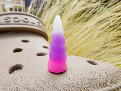 A tan shoes shoe decorated with several colorful unicorn horn charms, each one blending from one pastel shade to another in shades of pink, yellow, blue, and purple. The shoe is displayed on a backdrop of light green grasses.