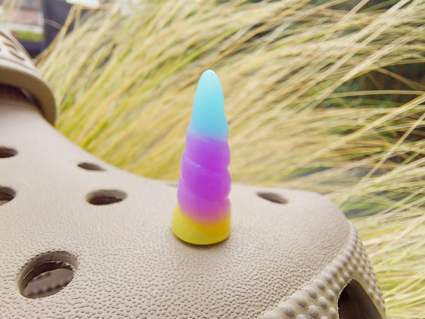 A tan shoes shoe decorated with several colorful unicorn horn charms, each one blending from one pastel shade to another in shades of pink, yellow, blue, and purple. The shoe is displayed on a backdrop of light green grasses.