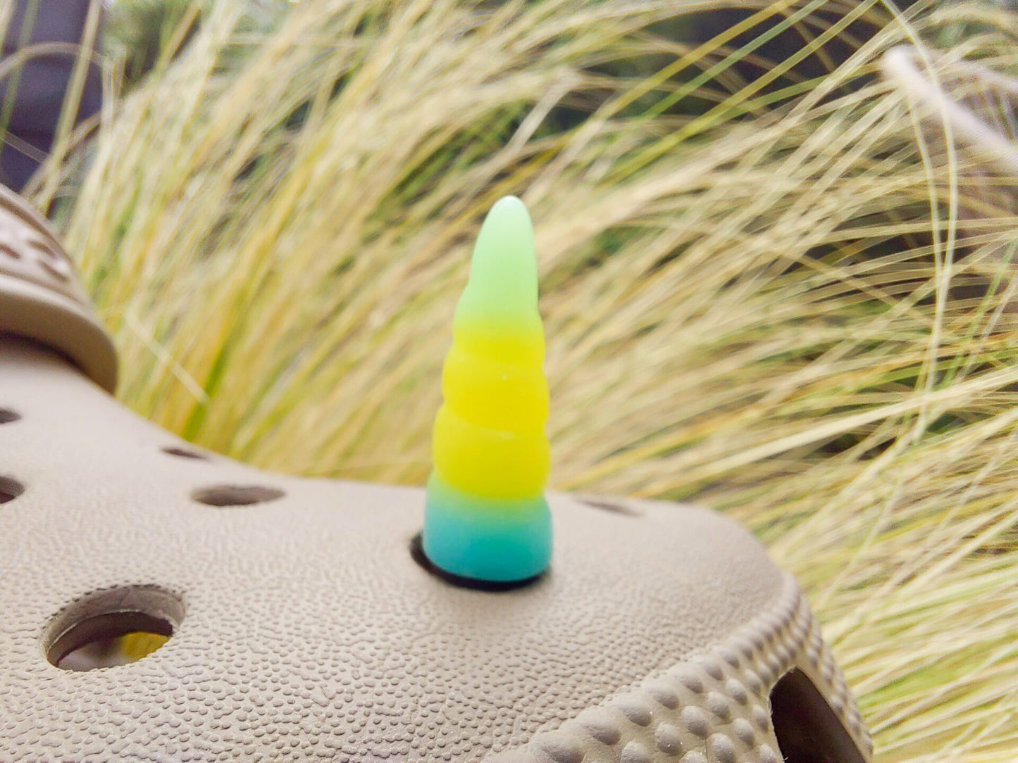 A tan shoes shoe decorated with several colorful unicorn horn charms, each one blending from one pastel shade to another in shades of pink, yellow, blue, and purple. The shoe is displayed on a backdrop of light green grasses.