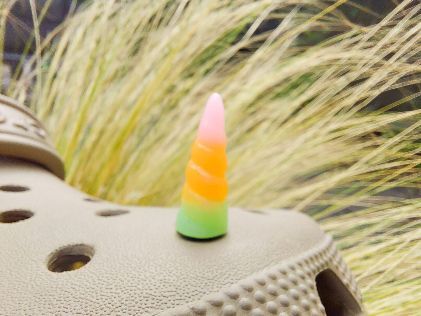 A tan shoes shoe decorated with several colorful unicorn horn charms, each one blending from one pastel shade to another in shades of pink, yellow, blue, and purple. The shoe is displayed on a backdrop of light green grasses.