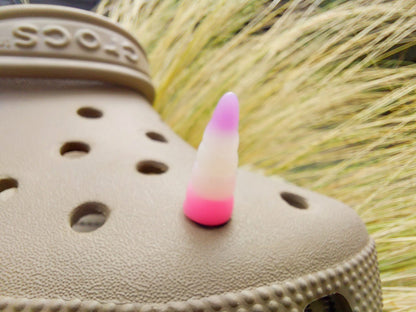 A tan shoes shoe decorated with several colorful unicorn horn charms, each one blending from one pastel shade to another in shades of pink, yellow, blue, and purple. The shoe is displayed on a backdrop of light green grasses.