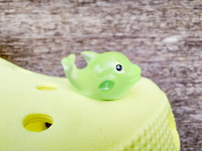 Six glow-in-the-dark dolphin charms in various pastel colors, including purple, pink, orange, green, blue, and white, are arranged on a yellow shoes shoe. The dolphins have tiny black eyes and wide grins, giving them a playful and cute appearance.