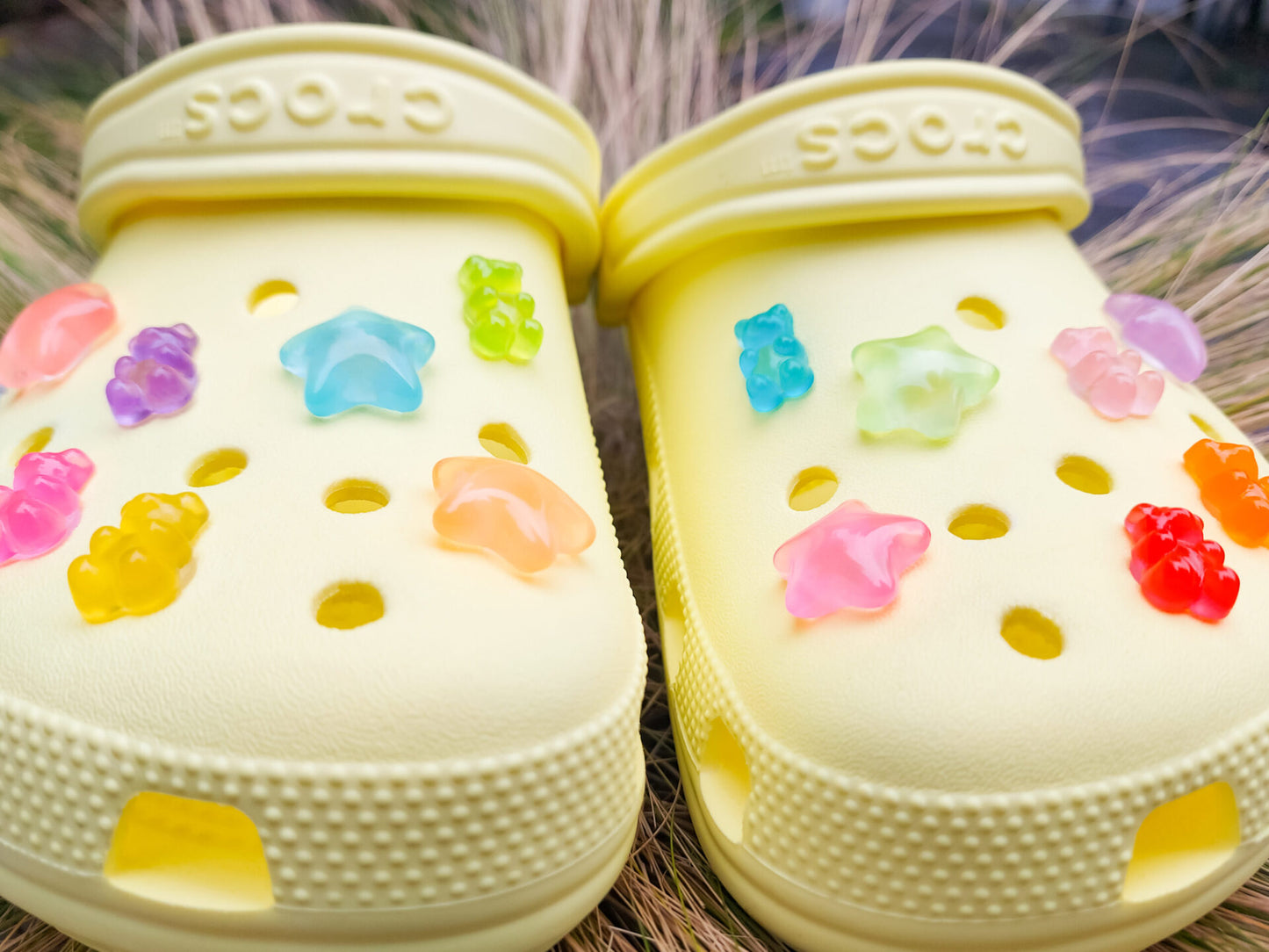 Close-up of yellow shoes featuring 14 colorful glow-in-the-dark gummy bear and star charms in shades of red, orange, pink, green, purple, and blue, arranged on the shoes, glowing softly in daylight against the bright background.