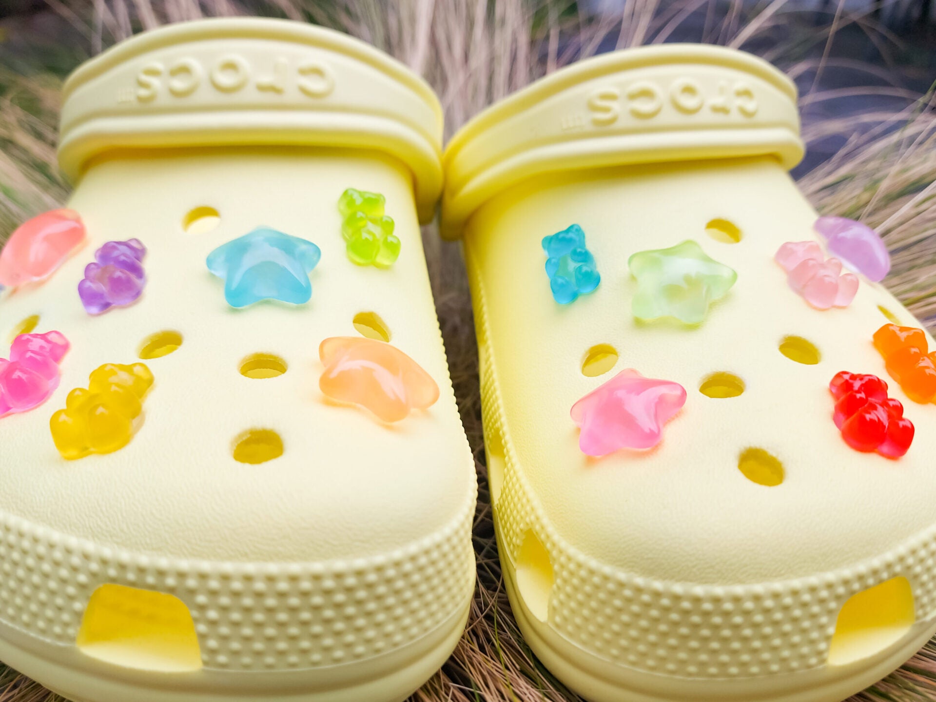 Close-up of yellow shoes featuring 14 colorful glow-in-the-dark gummy bear and star charms in shades of red, orange, pink, green, purple, and blue, arranged on the shoes, glowing softly in daylight against the bright background.