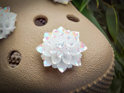 A pair of shoes adorned with multiple white rose charms featuring iridescent accents, placed on a background of green palm leaves. The charms add an elegant and sophisticated touch to the shoes.