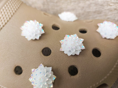 A pair of shoes adorned with multiple white rose charms featuring iridescent accents, placed on a background of green palm leaves. The charms add an elegant and sophisticated touch to the shoes.