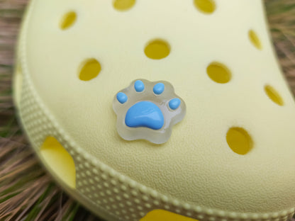 Glow-in-the-dark pastel paw print charms in pink, yellow, blue, and green, attached to a yellow shoe. The charms have a soft glow effect and add a playful animal-inspired look.